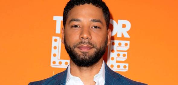 Jussie Smollett stares at the camera while wearing a white shirt and blue jacket as he stands in front of an orange background