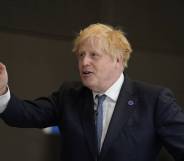 Boris Johnson wears a dark suit jacket, white shirt and blue tie as he gestures to the side