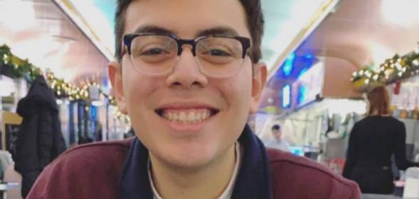 Julio Ramirez is seen smiling at the camera while wearing a black and red jacket with a white shirt and wearing black rimmed glasses