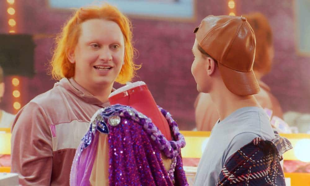 Jinkx Monsoon stands next to a dress form with purple fabrics on it as they talk to Trinity the Tuck, who is facing away from the camera