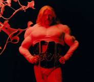 Tom Rasmussen wears a white top and black corset as they stand with their hands on their hips and are lit by a red light