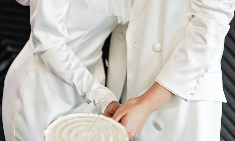 Two people, who appear to be female presenting, wear white wedding clothes including a dress and suit while cutting a white cake and holding hands