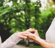 Two female presenting people, who are presumed to be in a lesbian or queer relationship, face each other while wearing wedding dresses as one person puts a ring on the other's hand