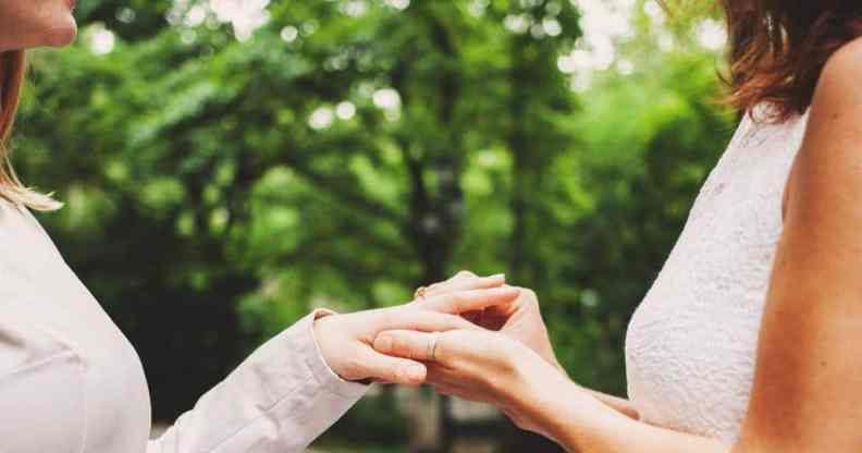 Two female presenting people, who are presumed to be in a lesbian or queer relationship, face each other while wearing wedding dresses as one person puts a ring on the other's hand