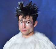 Ezra Miller wears a white fluffy top with their hair spiked up and wearing silver makeup on their face