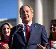 Texas attorney general Ken Paxton wears a white button up shirt, red tie and blue suit jacket as he speaks into a microphone and gestures with both his hands. He is standing in front of two people wearing red outfits