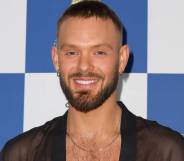 John Whaite smiles at the camera while wearing a black mesh top with a gold necklace as he stands in front of a white and blue checkered background