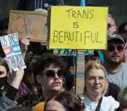 Several people gather in a crowd in support of the trans community in the UK. One person holds up a sign reading 'trans is beautiful' in the crowd.