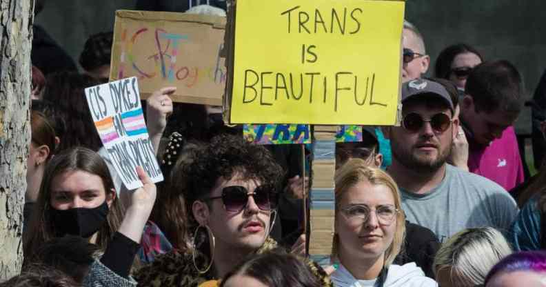 Several people gather in a crowd in support of the trans community in the UK. One person holds up a sign reading 'trans is beautiful' in the crowd.