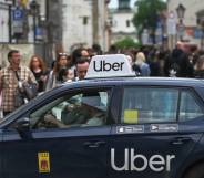 A person stands outside a black car that has a white Uber logo on the back door as well as a white Uber sign on the roof