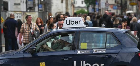 A person stands outside a black car that has a white Uber logo on the back door as well as a white Uber sign on the roof