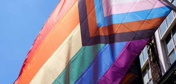 The progressive Pride flag flies outside a building, against a bright blue sky.