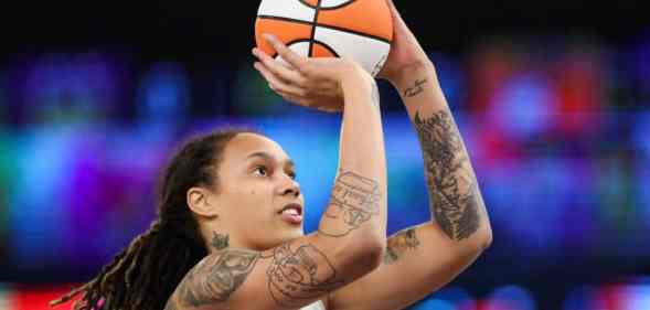 Brittney Griner wears a white jersey as she holds up a white and orange basketball as she shoots it towards a hoop. Her hair is styled in locks and she has tattoos on her arms