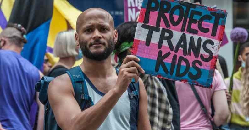 A person holds up a sign coloured in blue, pink and white like the trans Pride flag which reads 'Protect trans kids'