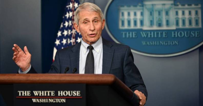 Anthony Fauci stands at a podium in the White House with the White House logo and an American flag in the background. Fauci is wearing a white shirt, dark tie and dark suit jacket as he talks to people off camera