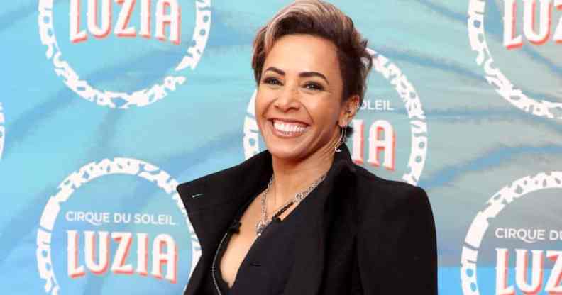 Dame Kelly Holmes wears a black top and black jacket as she smiles at the camera and stands in front of a blue background