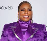 Aunjanue Ellis smiles at the camera while wearing a bright purple turtleneck top and matching jacket with silver lining on the lapel. She is wearing gold dangle earrings and pinky-purple eyeshadow to match her clothing