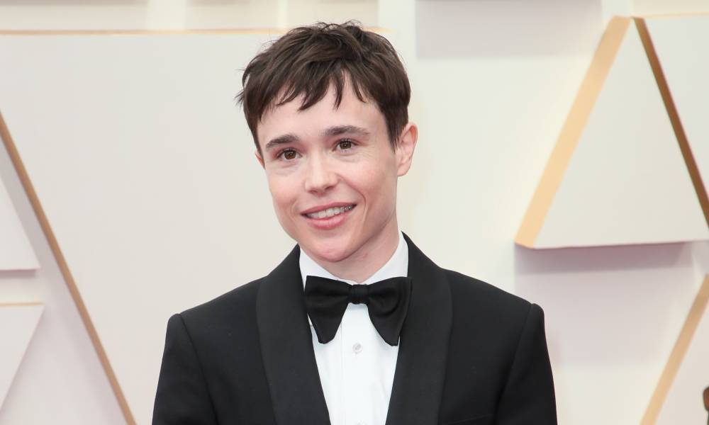 Elliot Page smiles for the camera while wearing a bow tie, white button up shirt and black suit jacket and standing in front of a pale off-white background