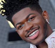 Lil Nas X smiles at the camera showing off decorative sparkly grills on his teeth. He has light blue eye shadow at the inner corner of his eyes and is wearing a while outfit with sparkly details on the collar and a matching dangly earring in his ear