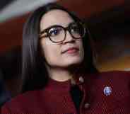 Alexandria Ocasio-Cortez, known as AOC, wears a red and black striped jacket with a dark top underneath with a little pin as she stares at something off camera