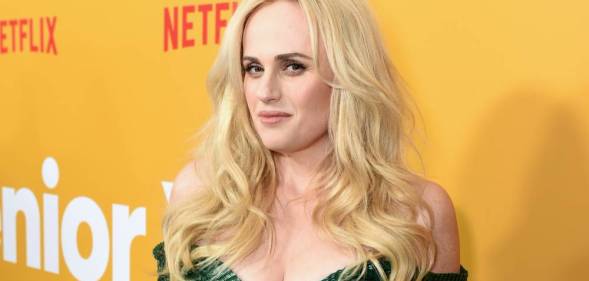 Rebel Wilson wears a green dress with her blonde hair down as she stands in front of a yellow background with the red Netflix logo in the background