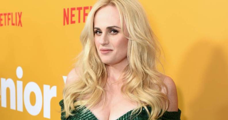 Rebel Wilson wears a green dress with her blonde hair down as she stands in front of a yellow background with the red Netflix logo in the background