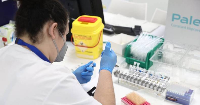 A scientist wearing a grey face mask, blue shirt and white top holds a dropper and container in their blue gloved hands as monkeypox samples are seen on their desk