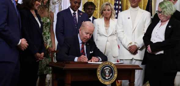 President Biden sits at a desk, signing an executive order, while LGBT+ advocates watch