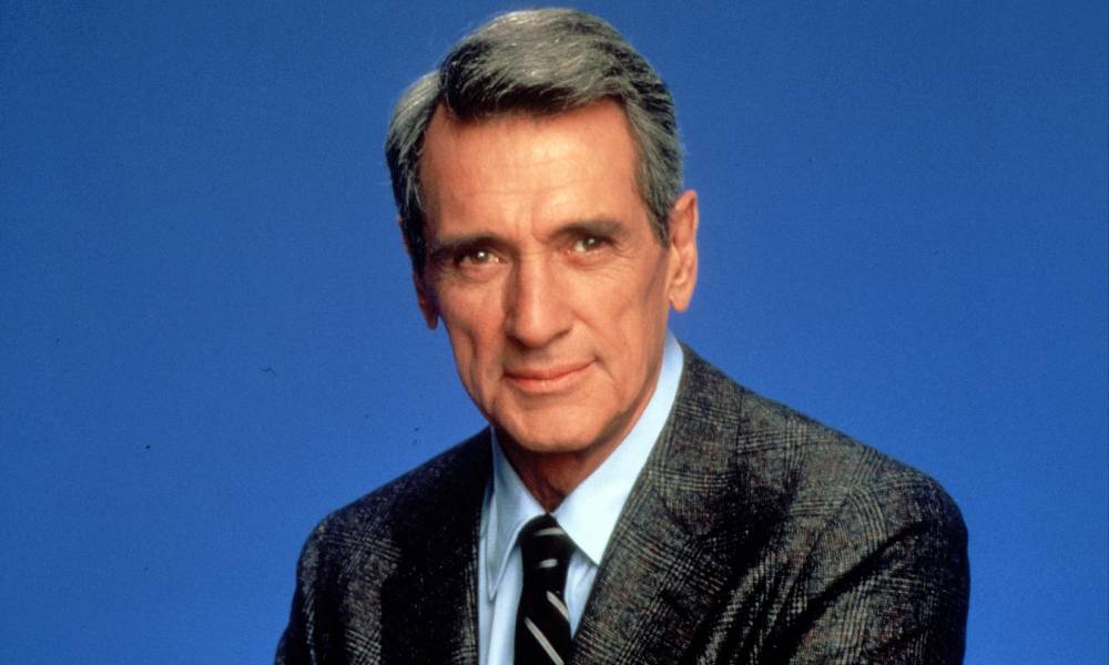 Rock Hudson stares at the camera while wearing a light blue button up, a striped tie and a grey suit jacket