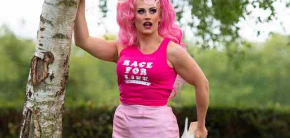 Drag Race UK star Ella Vaday wears a pink wig, hot pink sleeveless shirt with a white 'Race for Life' logo on it and baby pink short skirt. She poses with one hand on a tree while holding one foot, which has a white high heel on it, in her other hand