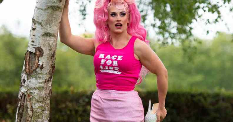 Drag Race UK star Ella Vaday wears a pink wig, hot pink sleeveless shirt with a white 'Race for Life' logo on it and baby pink short skirt. She poses with one hand on a tree while holding one foot, which has a white high heel on it, in her other hand