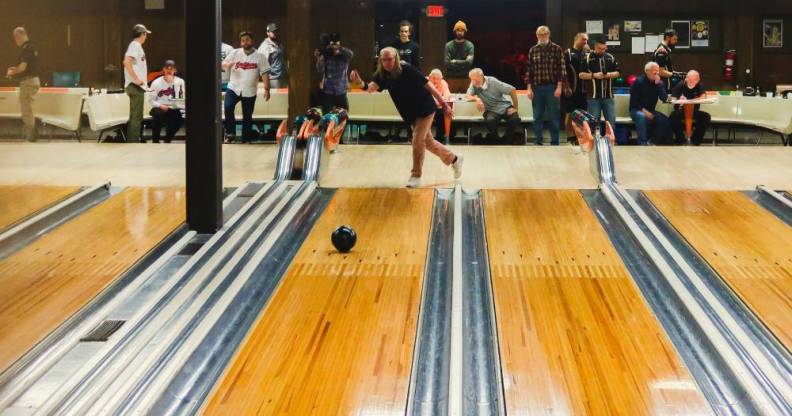 Michelle Guzowski throws a bowling ball down the lane of a bowling alley while other people in the background watch her bowl