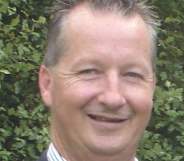 A headshot of Peter Keeley smiling in front of a bush