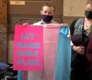 A person wearing a black face mask holds up a trans Pride flag and a sign reading "Let trans girls play" in protest of trans sports bans being passed