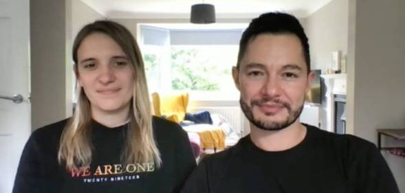 Hannah and Jake Graf sit in their house while wearing black shirts. Hannah wears a shirt that says 'we are one' in rainbow font while Jake sits next to her also wearing a black shirt. In the background, there are pillows and blankets on a yellow chair