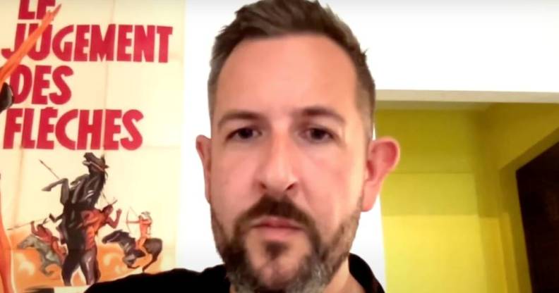 James McFadzean sits in a room wearing a dark shirt. The wall behind him is white and there is a poster with a person rising a bucking horse with words in French written on it