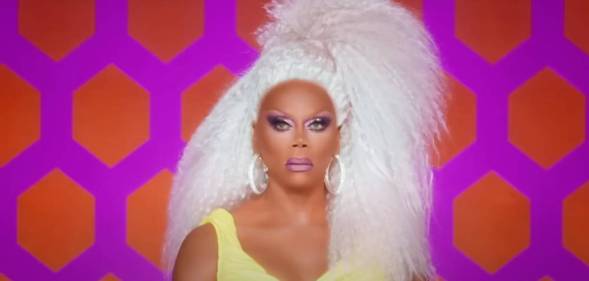 RuPaul wears a white blonde wig and yellow dress as the drag queen sits in front of a pink and orange patterned background