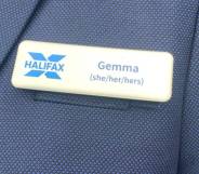 A Halifax employee wears a blue blazer and name badge which includes the name 'Gemma' and her pronouns 'she/her/hers' as well as the blue Halifax logo