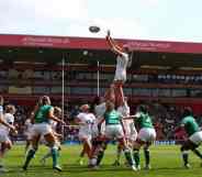 Several women wearing green or white rugby uniforms surround one player who is being held up by their teammates to catch the ball which is flying in the air towards them