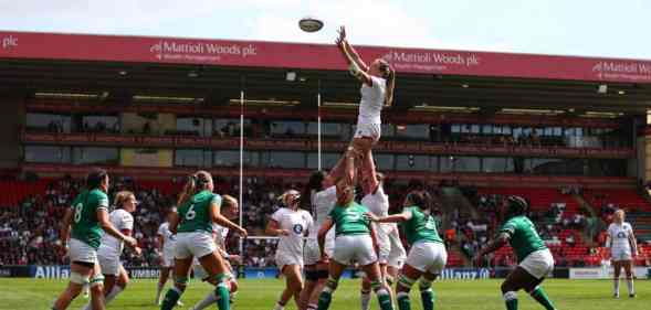 Several women wearing green or white rugby uniforms surround one player who is being held up by their teammates to catch the ball which is flying in the air towards them