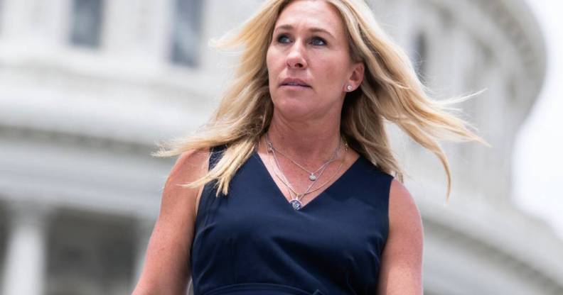 Marjorie Taylor Greene wears a dark blue dress as she stares off camera with her blonde hair blowing in the background. There is a white building seen in the background