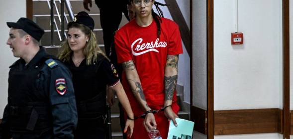 Brittney Griner wears a red short sleeved t-shirt with white writing on it as she is led down a stairway in Russia in handcuffs