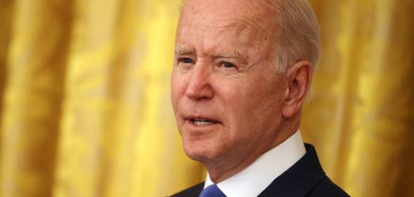 President Joe Biden is seen wearing a white button up shirt, blue tie and dark suit jacket as he stands in front of a yellow background