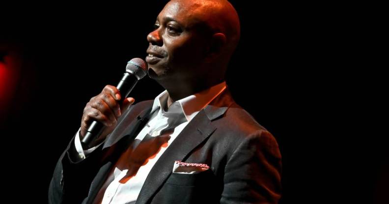 Dave Chappelle wears a white button up shirt, dark suit jacket and has something sticking out of the jacket's breast pocket as he speaks into a microphone he is holding is his hand