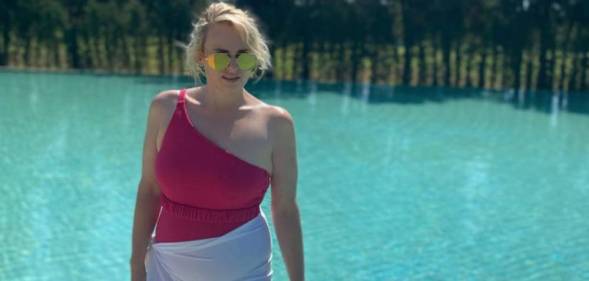 Rebel Wilson wears a pink off the shoulder swimsuit with a white cloth around her hips as she stands near a pool