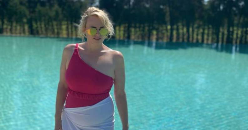 Rebel Wilson wears a pink off the shoulder swimsuit with a white cloth around her hips as she stands near a pool