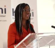 Kemi Badenoch stands at a transparent podium as she speaks to a crowd gathered off screen. She is wearing a red-orange dress with her hair styled in braids with white signs seen in the background.