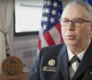 Dr Rachel Levine speaks to someone off camera while she is wearing a white shirt, dark jacket and pins denoting her status as a four-star admiral. There is a US flag seen in the background