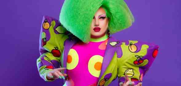 Drag Race UK star Choriza May wears a green wig, colourful outfit with large shoulder pads as she stands in front of a purple background