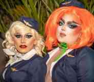 Two drag performers stand side by side, one with blonde hair and another with orange hair, as they appear to wear what looks like air cabin crew uniforms